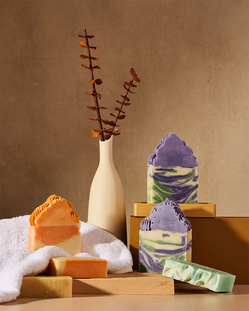 Royalty Soaps cold process handmade soap bars in purple, green, orange and cream with decorative soap frosting, displayed with a ceramic vase and eucalyptus on a warm neutral background.