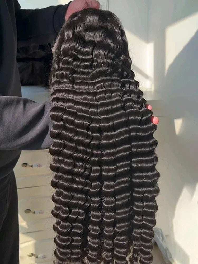 A person holding up a long, dark brown to black wavy hair extension or wig, showing the texture and volume of the curls. The background is a brightly lit room with white walls and light-colored furniture. 250 Density Black 42 Inch Human Hair Loose Deep Wave Curly 13x4 HD Lace Frontal Wig 13x6 Lace Front Wigs Clearance Sale Natural