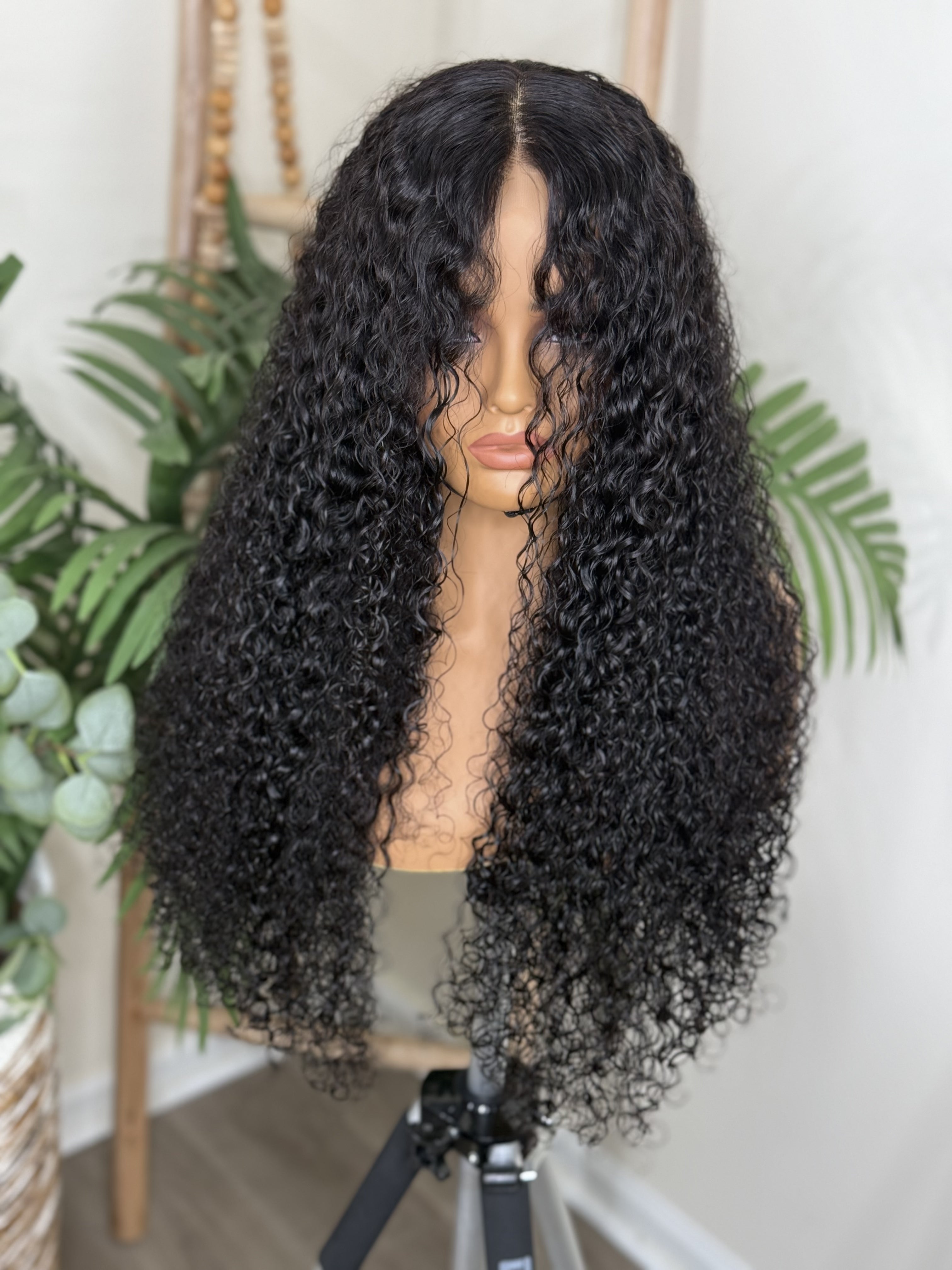 Black curly wig on a mannequin head with a white background and plants. Burmese Curly Virgin Hair - 100% Virgin Human Hair – Soft, full, and beautifully textured HD Lace Wig Glueless Wig