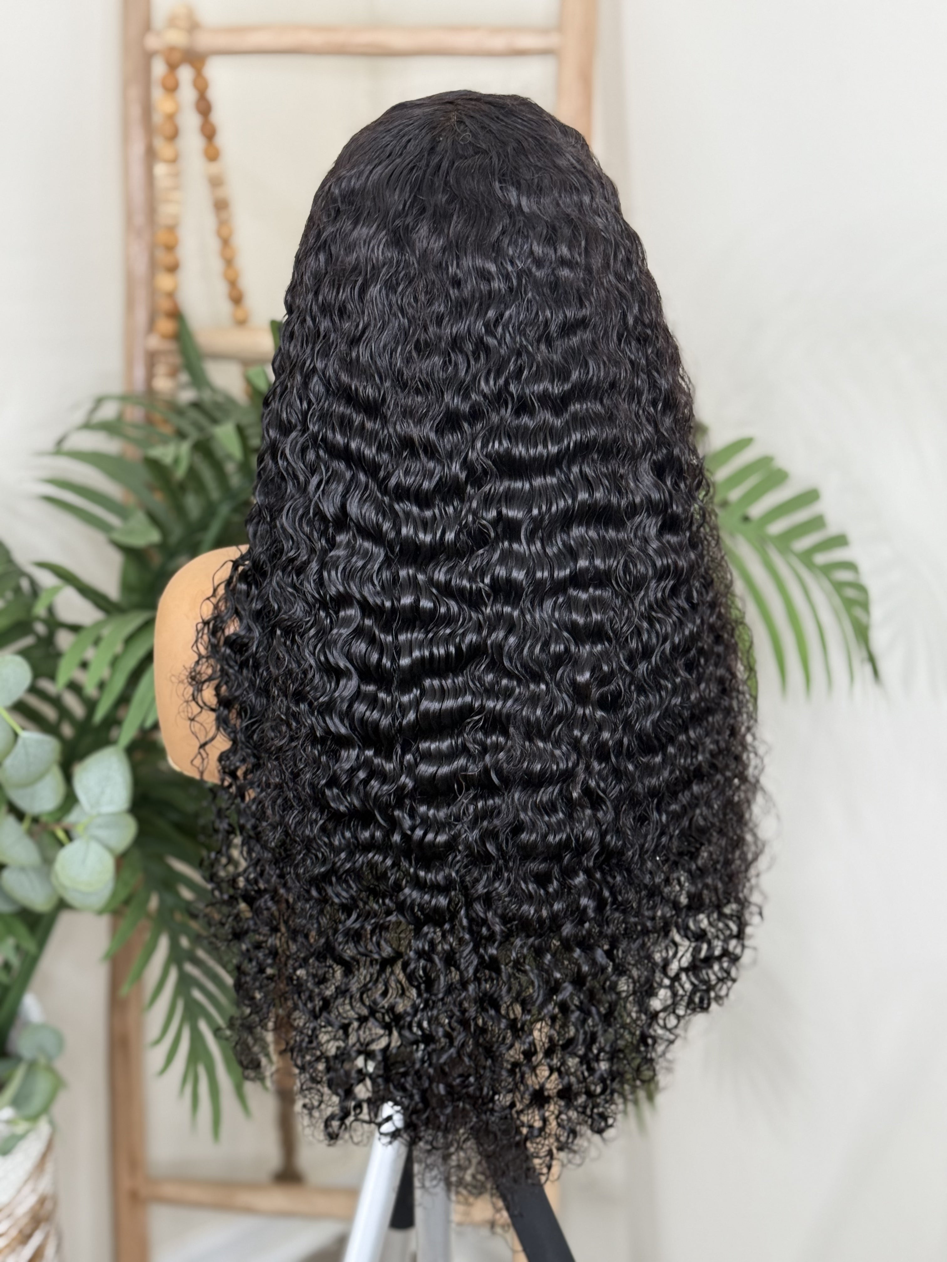 Black curly wig on a mannequin head with a white background and plants. Burmese Curly Virgin Hair - 100% Virgin Human Hair – Soft, full, and beautifully textured HD Lace Wig Glueless Wig