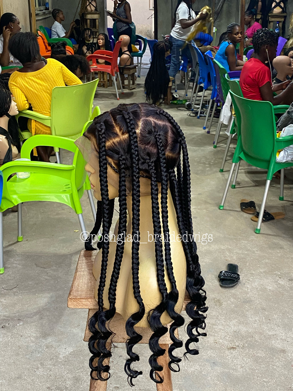 Person with styled hair in a salon setting with chairs and people in the background. jumbo knotless braid wig, knotless braid wig, jumbo braid wig, braided wig, full lace braided wig, swiss lace wig, swiss full lace wig, glueless braided wig, glueless wig, human hair braided wig, 100% human hair wig, knotless braids, jumbo knotless braids, scalp looking wig, natural scalp wig, lace braid wig, handmade braided wig, handcrafted wig, black braided wig, color 1B wig, 1B braided wig, long braided wig