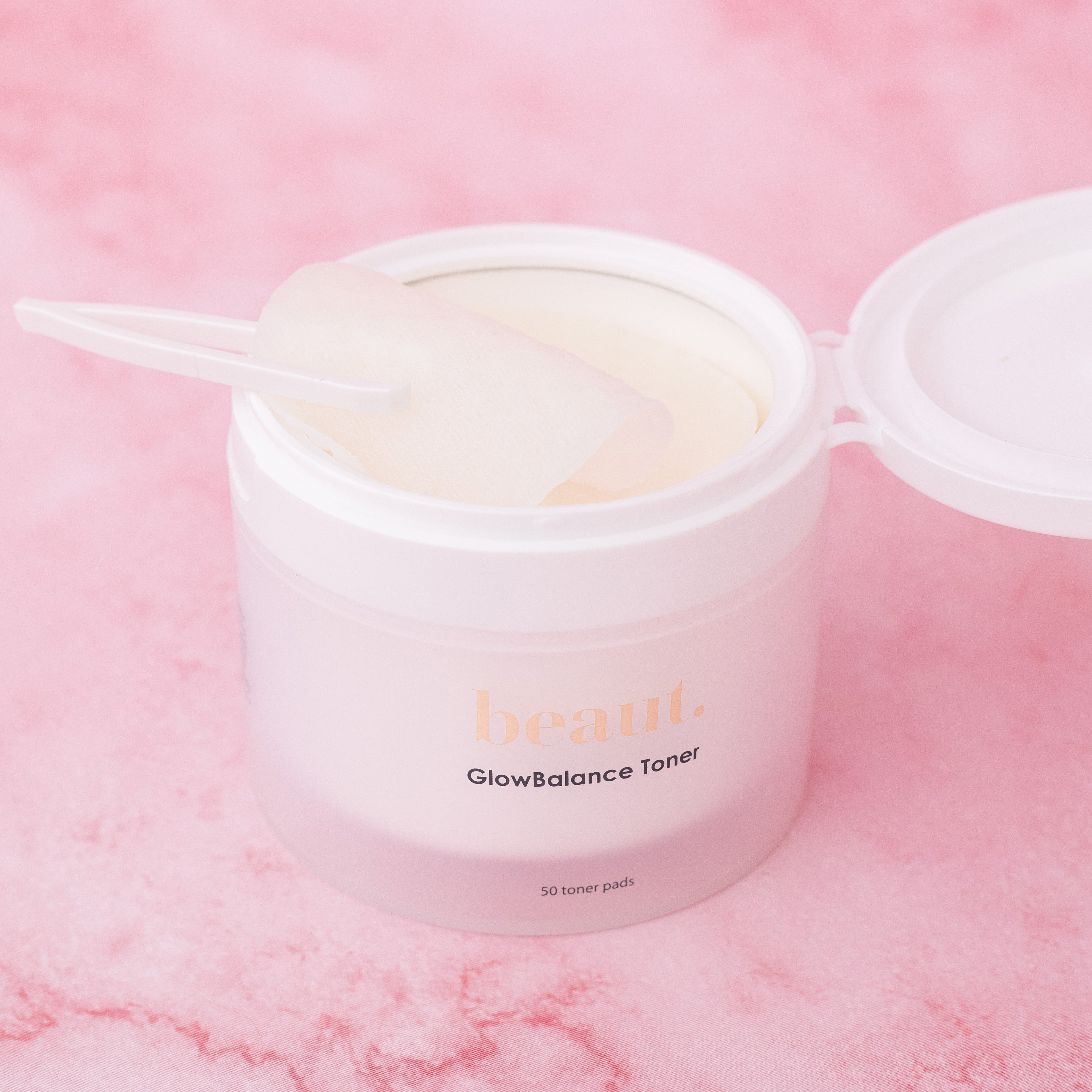 Beaut. GlowBalance Toner Pads with glycolic and lactic acid exfoliating facial toner pads, open 50-count skincare jar with toner pad applicator for radiant, smooth skin on pink background
