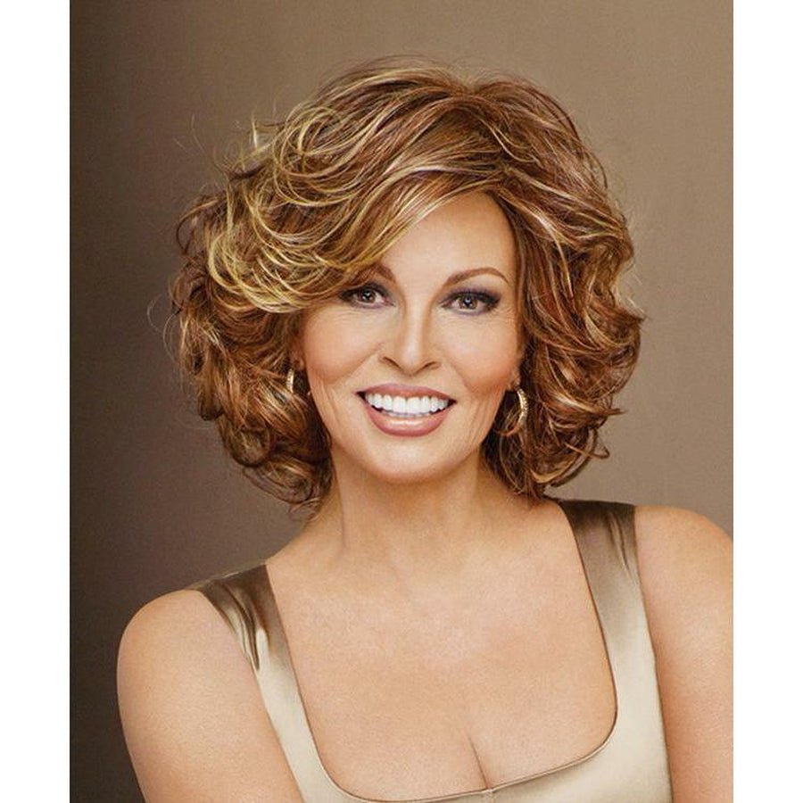 A smiling woman with a short, layered, curly wig in warm brown and golden blonde tones, wearing a beige top against a plain taupe background.