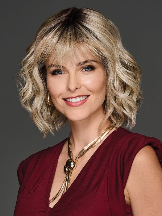 Woman with styled blonde hair wearing a red top and gold necklace on a gray background