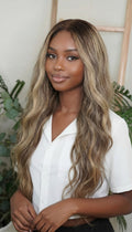 Woman with long, wavy hair wearing a white shirt in a natural setting. Honey Blonde Balayage Wave Wig 100% Virgin Human Hair – 22" Soft waves with sun-kissed dimension.