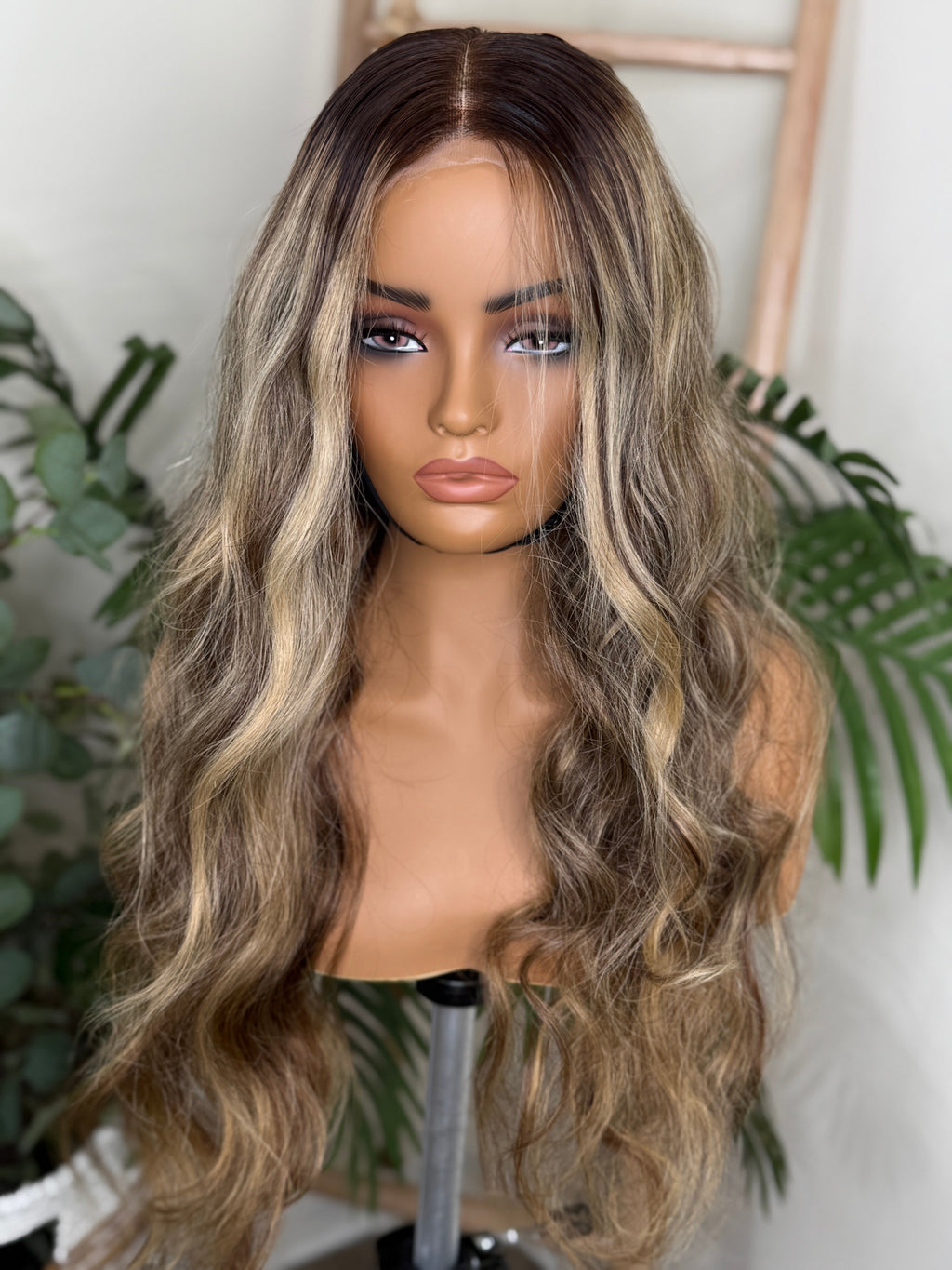 Mannequin head with long, wavy brown wig against a white background with green plants. Honey Blonde Balayage Wave Wig 100% Virgin Human Hair – 22" Soft waves with sun-kissed dimension.