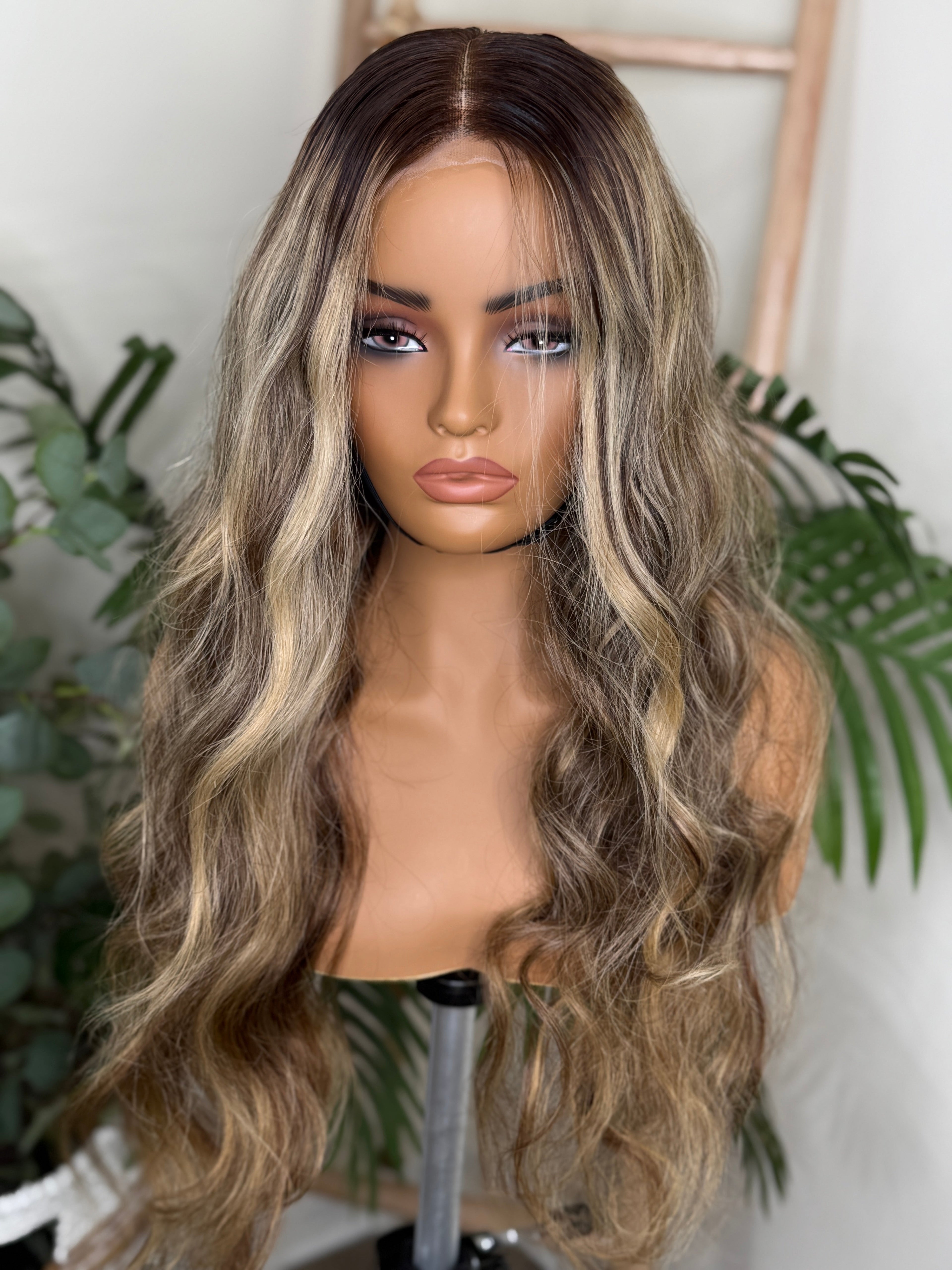 Mannequin head with long, wavy brown wig against a white background with green plants. Honey Blonde Balayage Wave Wig 100% Virgin Human Hair – 22" Soft waves with sun-kissed dimension.