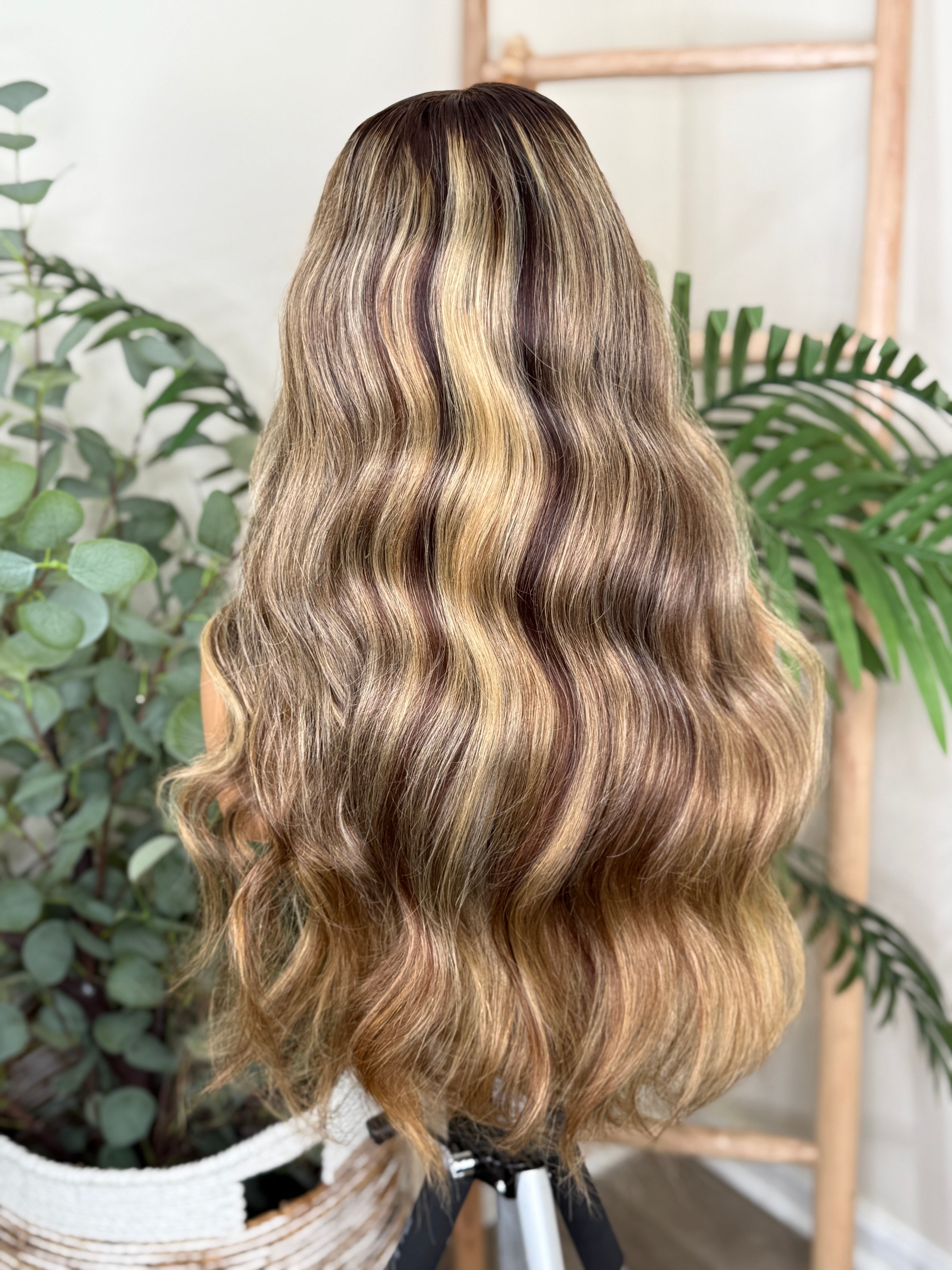 Person with wavy, ombre hair styled in a ponytail against a plant-filled background Honey Blonde Balayage Wave Wig 100% Virgin Human Hair – 22" Soft waves with sun-kissed dimension.