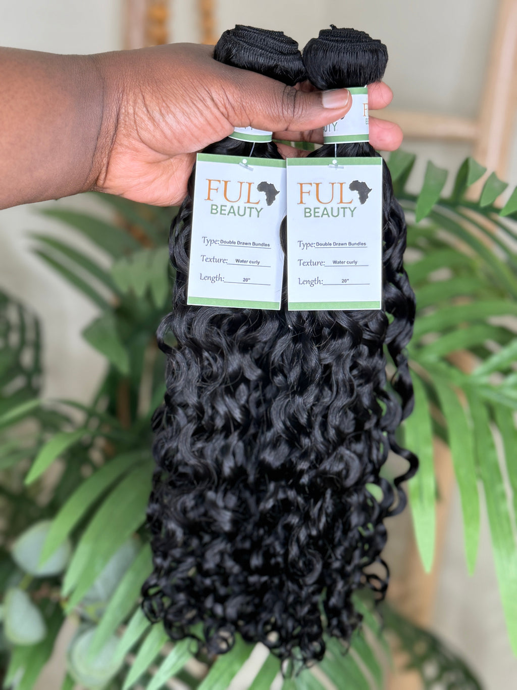 Hand holding two bundles of black hair extensions with 'FUL Beauty' labels against a blurred green plant background. Water Curly Wefted Bundle - 100% Human Hair, Double Drawn 18" to 26" inch Bundles