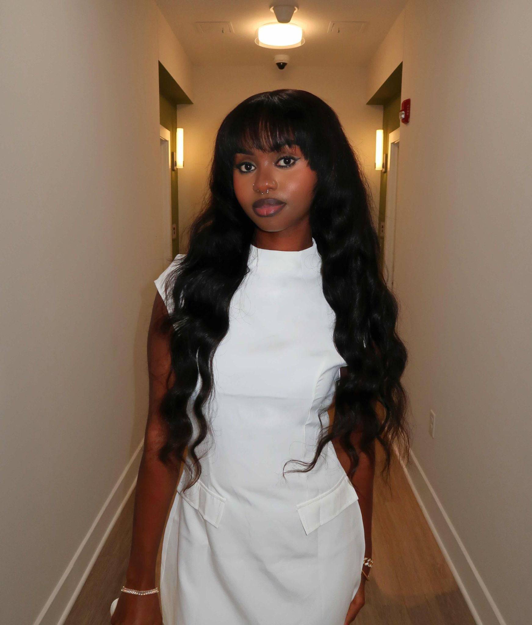 Woman with long, wavy hair wearing a white outfit in a hallway. Body Wave 100% Virgin Human Hair – No tangles, no shedding, all softness. 16”–30”