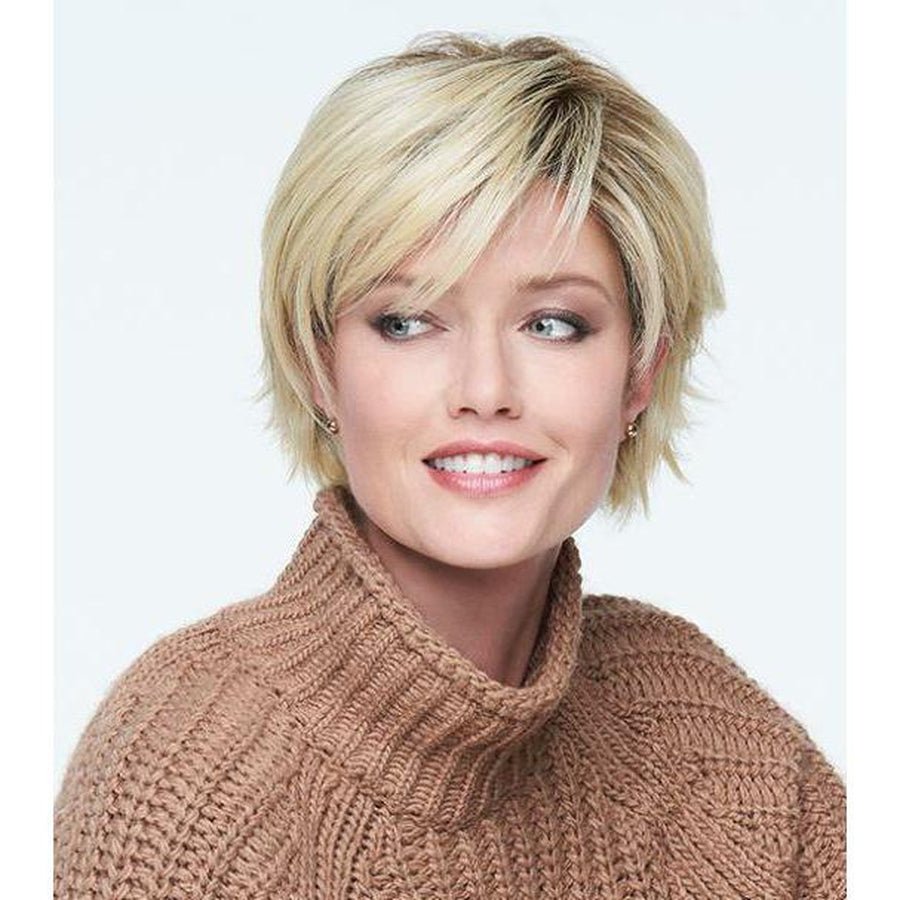 A woman with a short blonde pixie cut wearing a brown cable-knit turtleneck sweater, shown from the chest up against a light gray studio background.