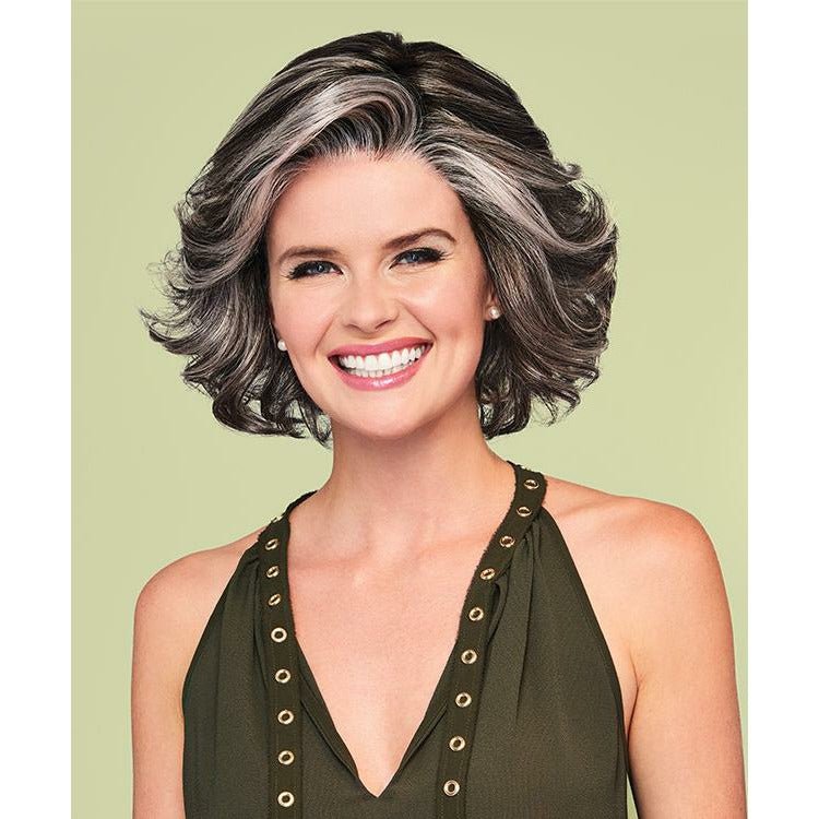 A smiling woman with a short, layered, wavy gray-brown wig is shown from the chest up against a solid pale green background. She wears an olive green halter top with gold-tone eyelet trim.
