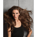 A woman with voluminous, wavy medium brown hair wearing a black top, posing against a textured gray concrete wall.