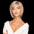 A woman modeling a short, straight, chin-length blonde wig with side-swept bangs against a solid black background.