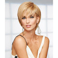 A woman modeling a short, layered blonde wig with side-swept bangs and geometric wire earrings against a bright, softly blurred window background.